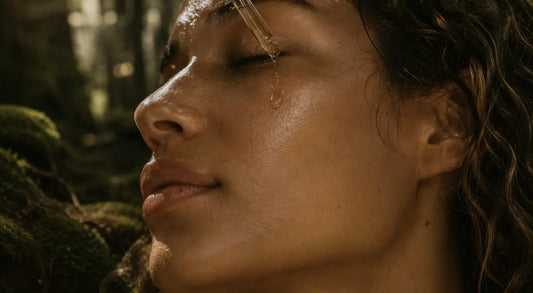 Woman with dewy skin in a misty pine forest as golden facial oil drips from a glass dropper onto her closed eyelid, surrounded by moss-covered rocks and soft shadowed woodland light.