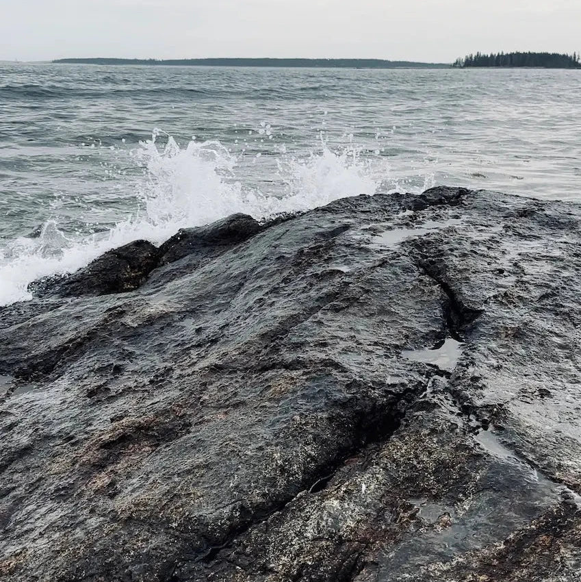A close-up view of dark, wet rocks in the foreground with ocean water splashing onto the shore under an overcast, cloudy sky.
