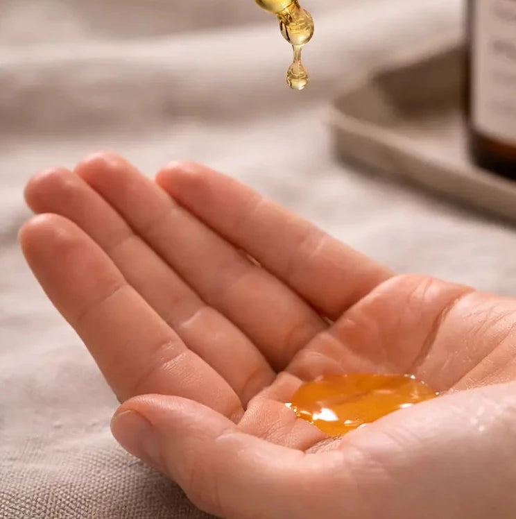 Hand holding a dropper with yellow liquid over another hand, with a bottle in the background.